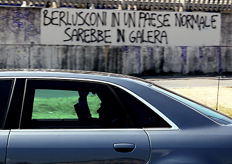 El primer ministro italiano pasa frente a un graffiti dirigido contra él. 'Berlusconi en un país normal estaría en prisión' se lee. | David Bebber