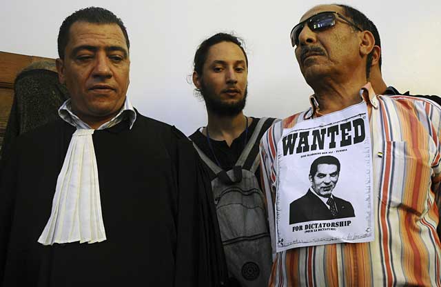 Un manifestante con una pancarta contra Ben Ali cerca de un abogado en el tribunal de Túnez que juzga al ex presidente. | AP